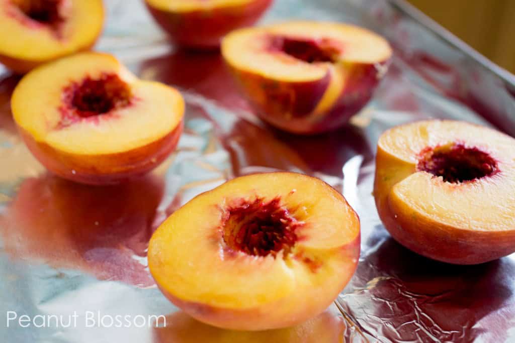 The peaches have been sliced in half and the pits removed. They are placed on a baking sheet cut side up, the baking sheet is covered in aluminum foil.