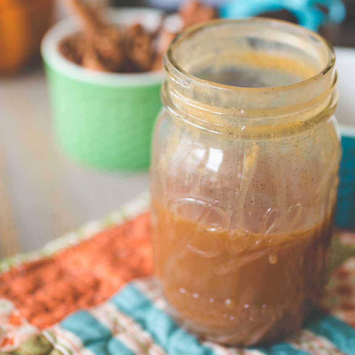 A jar of homemade caramel sauce sits on a table runner next to a bowl of cinnamon sticks.