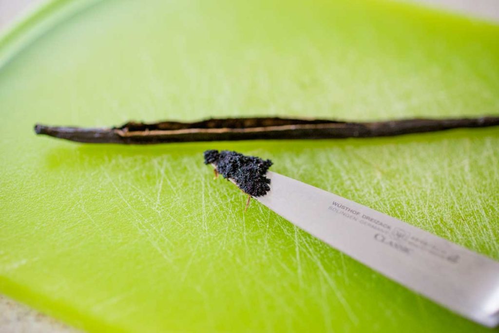A fresh vanilla bean pod has been sliced open and a knife is showing the vanilla seeds.
