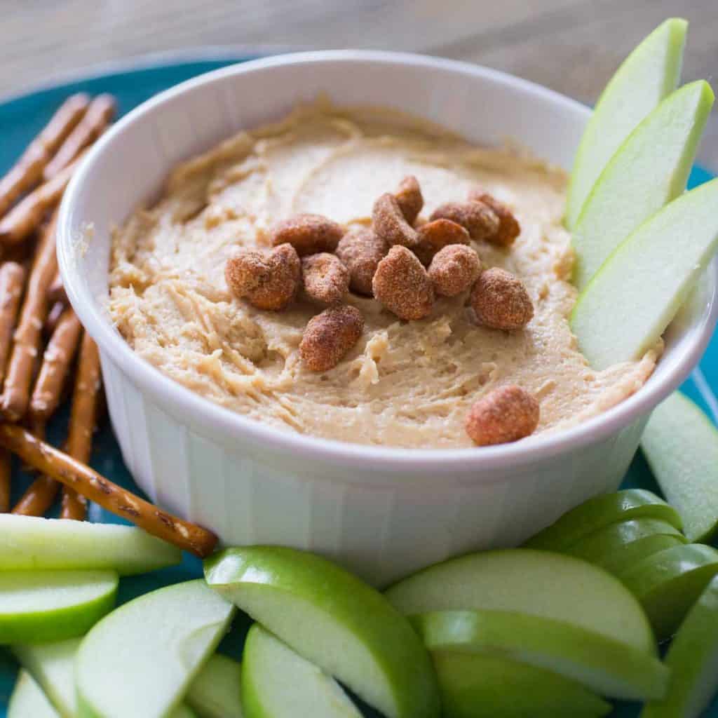 A serving bowl has creamy peanut butter apple dip and is topped with salted peanuts. There are green apple slices in the dip and on the plate with pretzels.