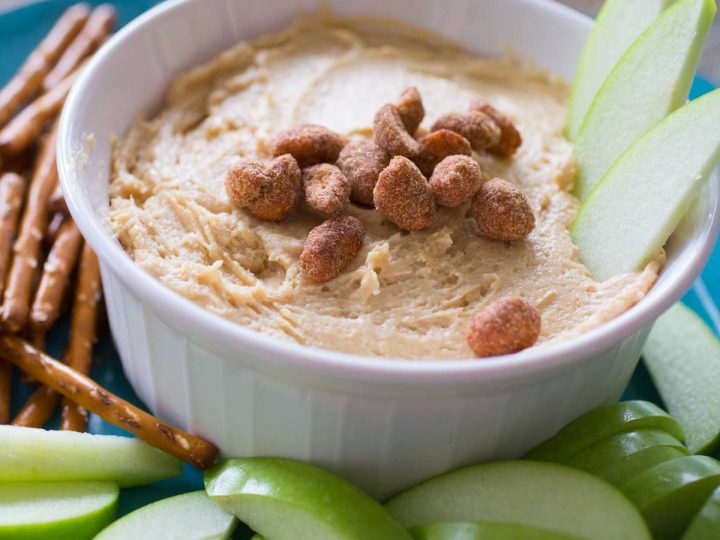 A serving bowl has creamy peanut butter apple dip and is topped with salted peanuts. There are green apple slices in the dip and on the plate with pretzels.