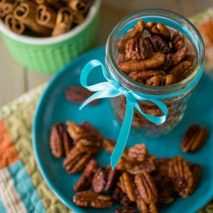 A mason jar filled with spiced pecans has a blue ribbon tied around it and sits on a blue plate with more pecans sprinkled around.