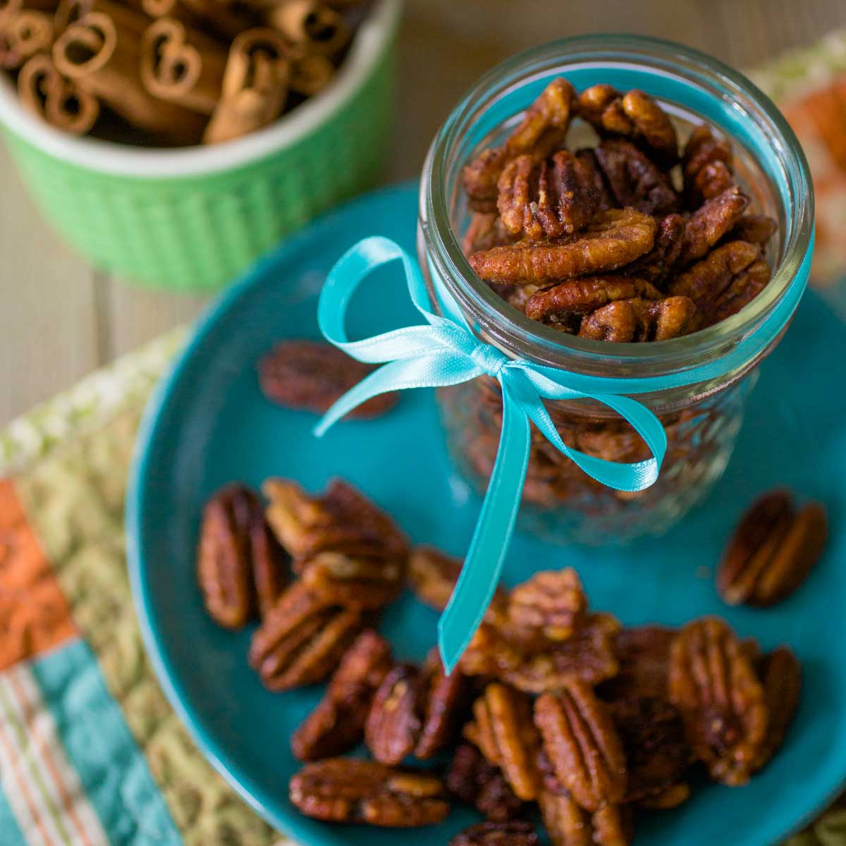 A mason jar filled with spiced pecans has a blue ribbon tied around it and sits on a blue plate with more pecans sprinkled around.