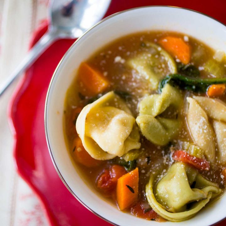 A bowl filled with tortellini soup shows the pasta, chunks of carrots, and a tomato based broth. A spoon sits next to the bowl.