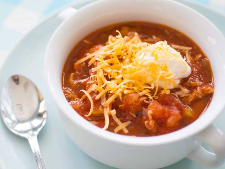 A white bowl of tomato-based chicken chili has shredded cheddar and a dollop of sour cream on top.