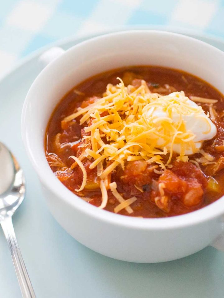 A white bowl of tomato-based chicken chili has shredded cheddar and a dollop of sour cream on top.