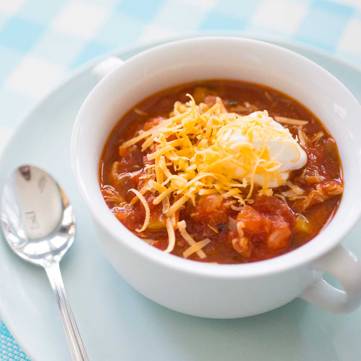A white bowl of tomato-based chicken chili has shredded cheddar and a dollop of sour cream on top.