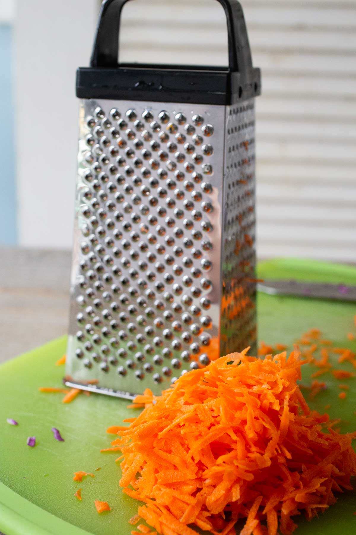 A box grater sits on a cutting board next to a pile of shredded carrots.