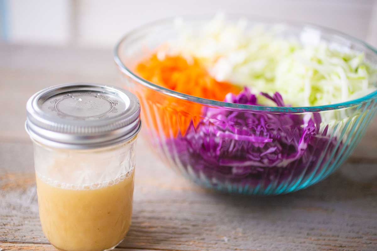A mason jar with homemade coleslaw dressing sits next to the mixing bowl of shredded vegetables.