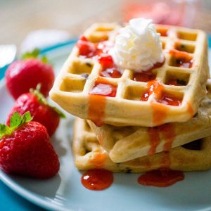 A stack of peanut butter waffles on a blue plate has strawberry jam running down the sides and a dollop of whipped cream on top.