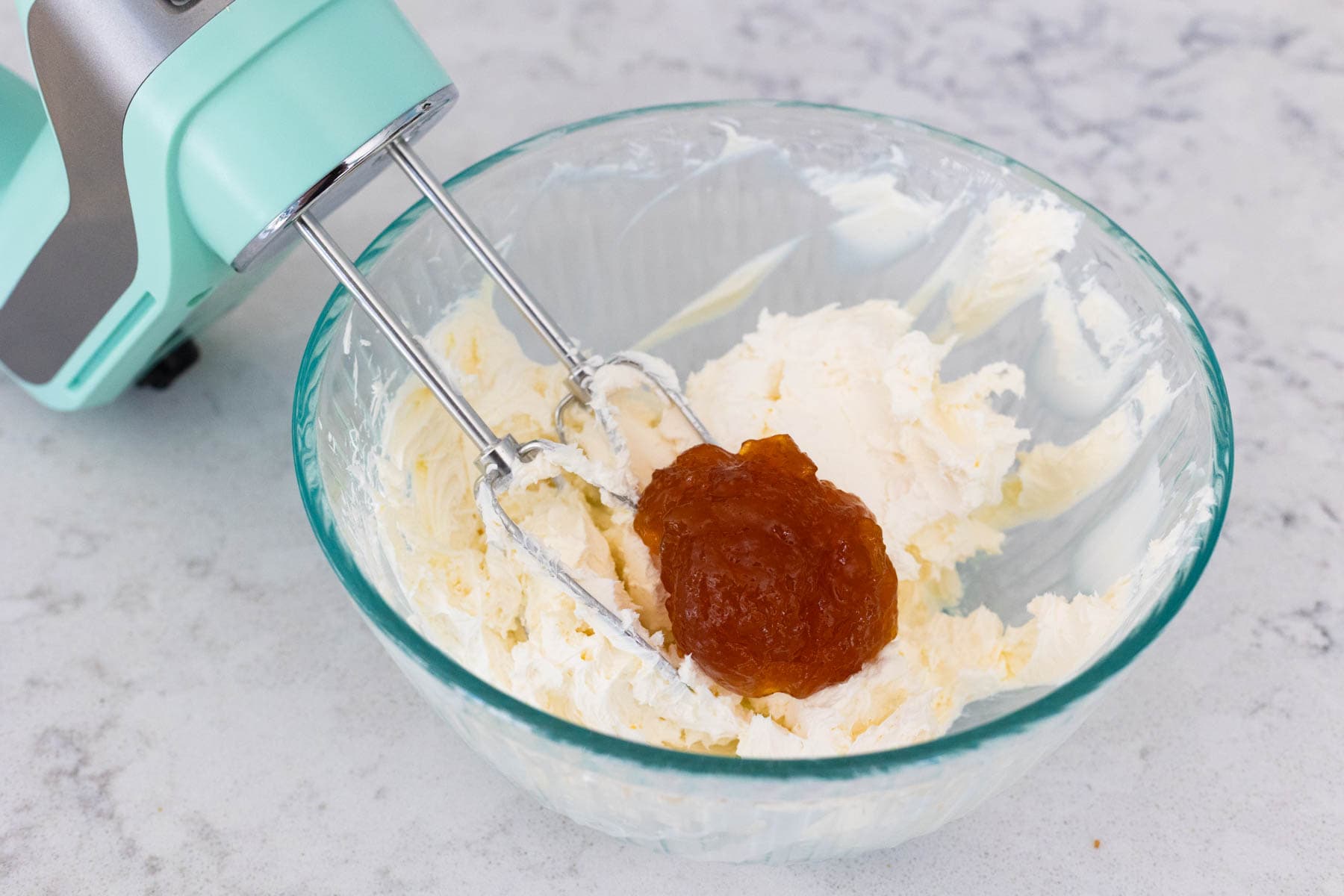 The mixing bowl has the hand mixer beating cream cheese and fruit preserves together.
