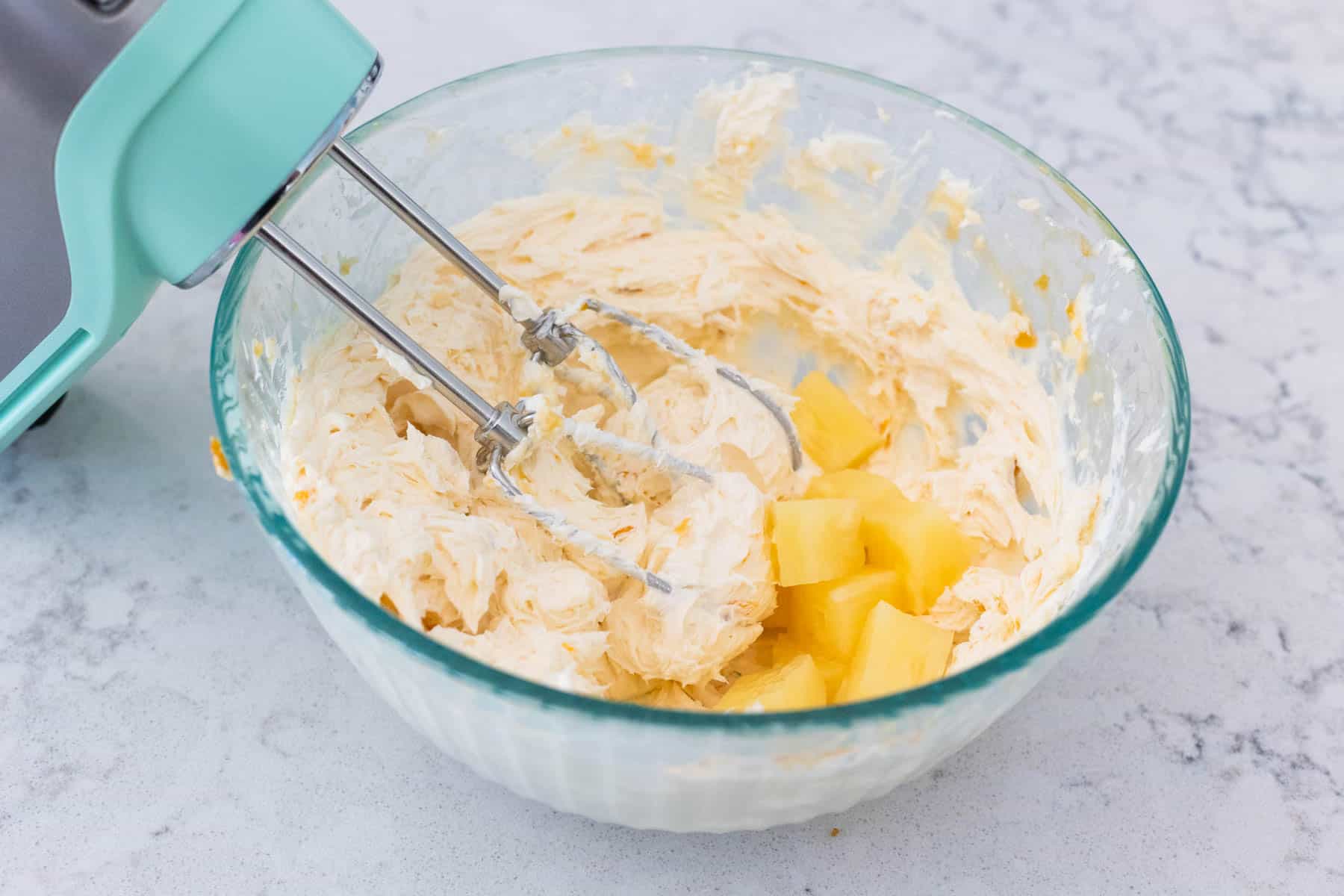 The pineapple chunks have been added to the bowl and will be crushed by the hand mixer blades.