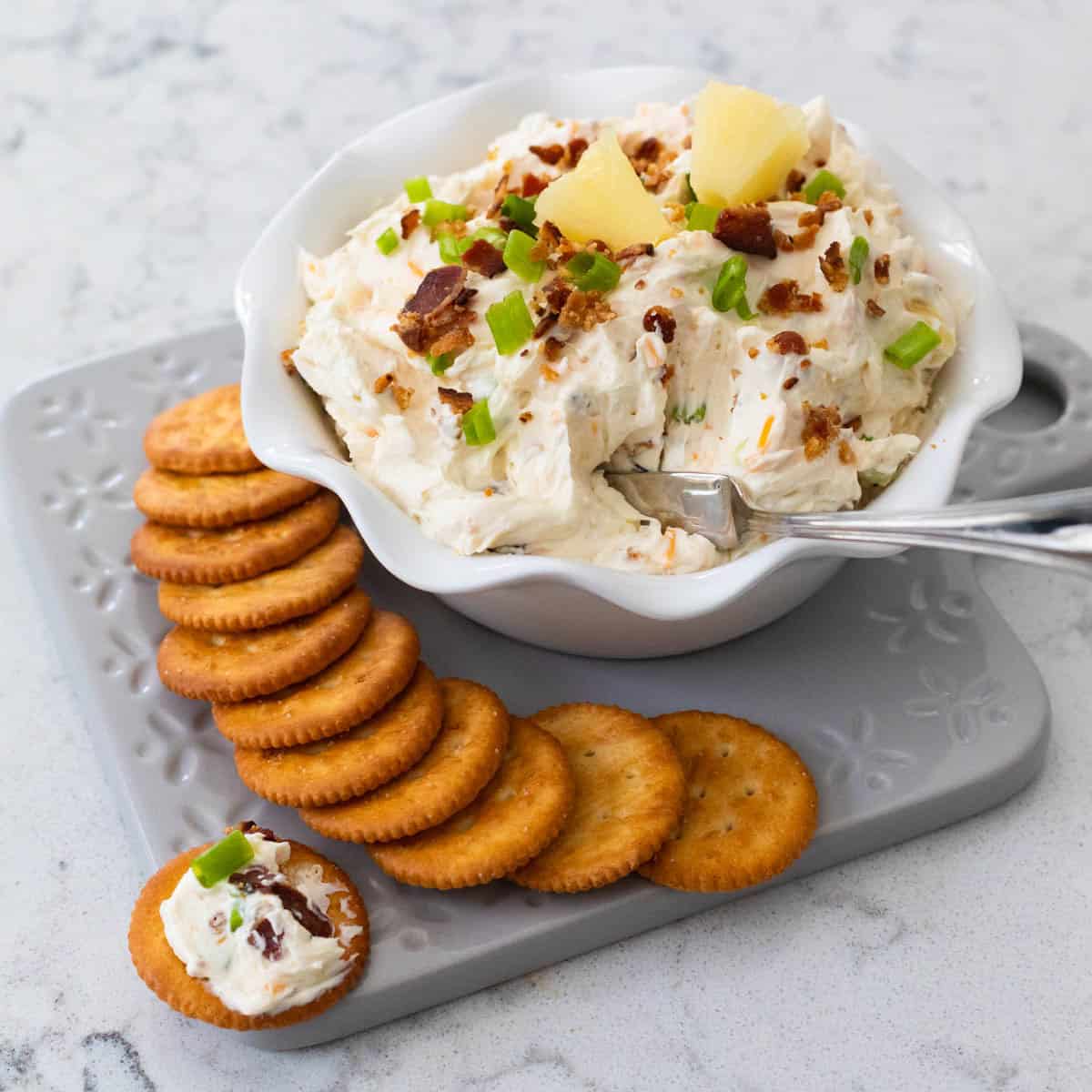 The bowl of creamy dip has bacon, green onions, and pineapple on top. A row of crackers are arranged on the platter next to it.