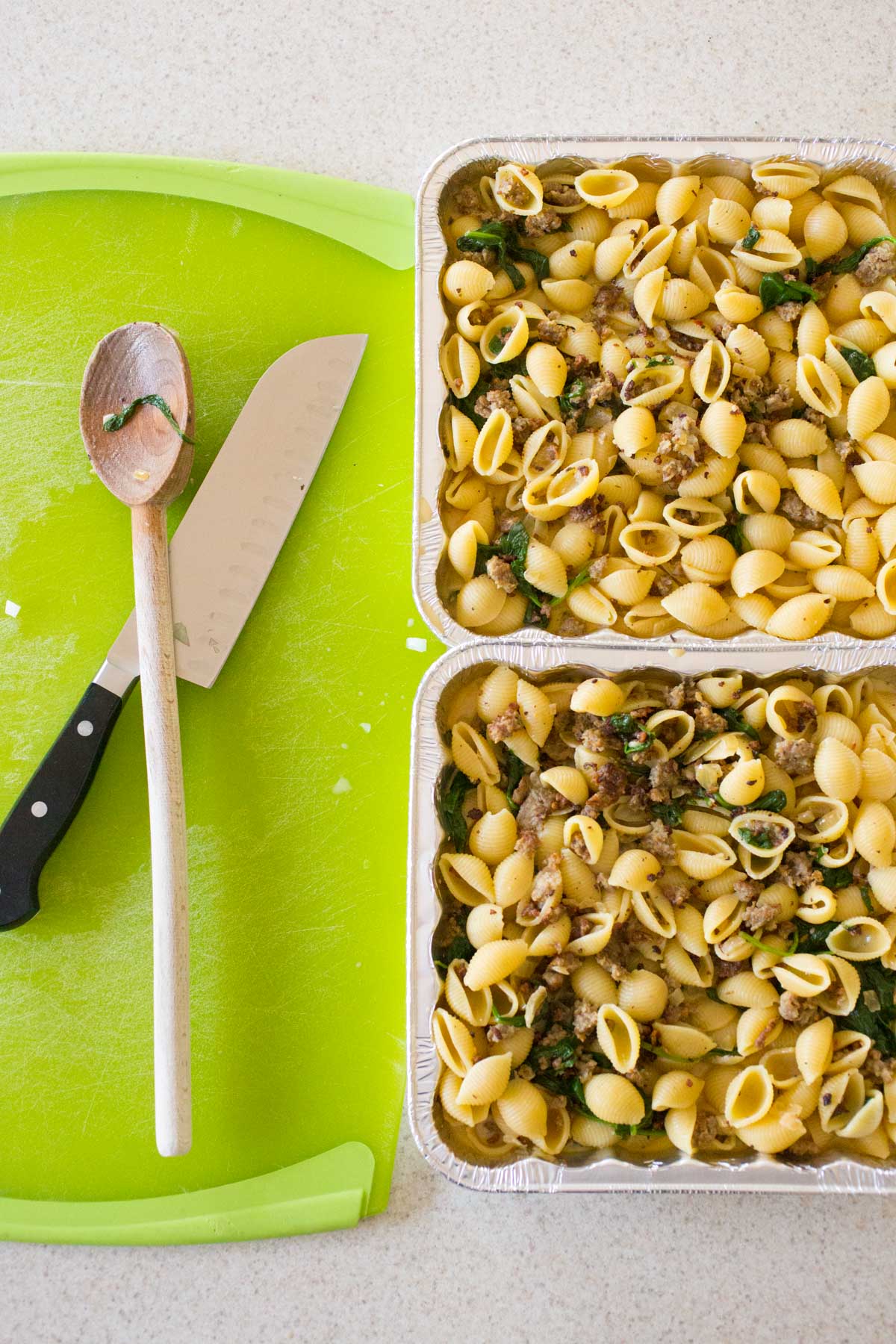 The pasta has been stirred into the mixture and placed in two 8x8-inch baking pans. A spoon and a knife rest on a cutting board next to them.