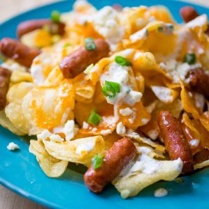 A plate of potato chips has buffalo and blue cheese dressing drizzled over he top with green onions sprinkled and little sausages scattered.