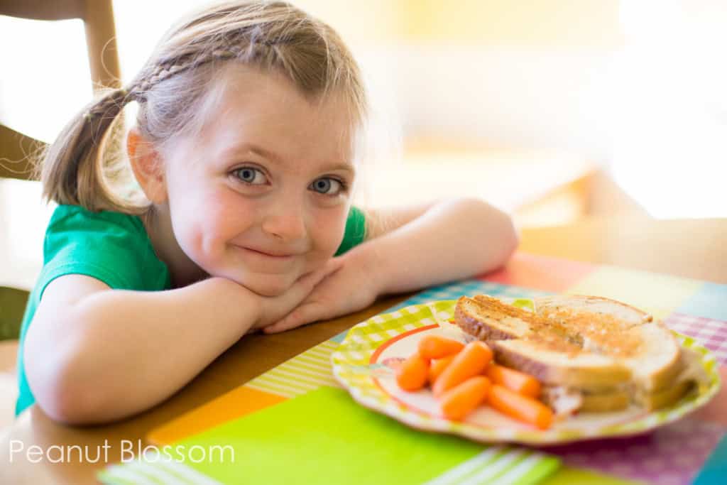 A young girl in a green shirt sits next to a dinner plate with a reuben sandwich and carrots.