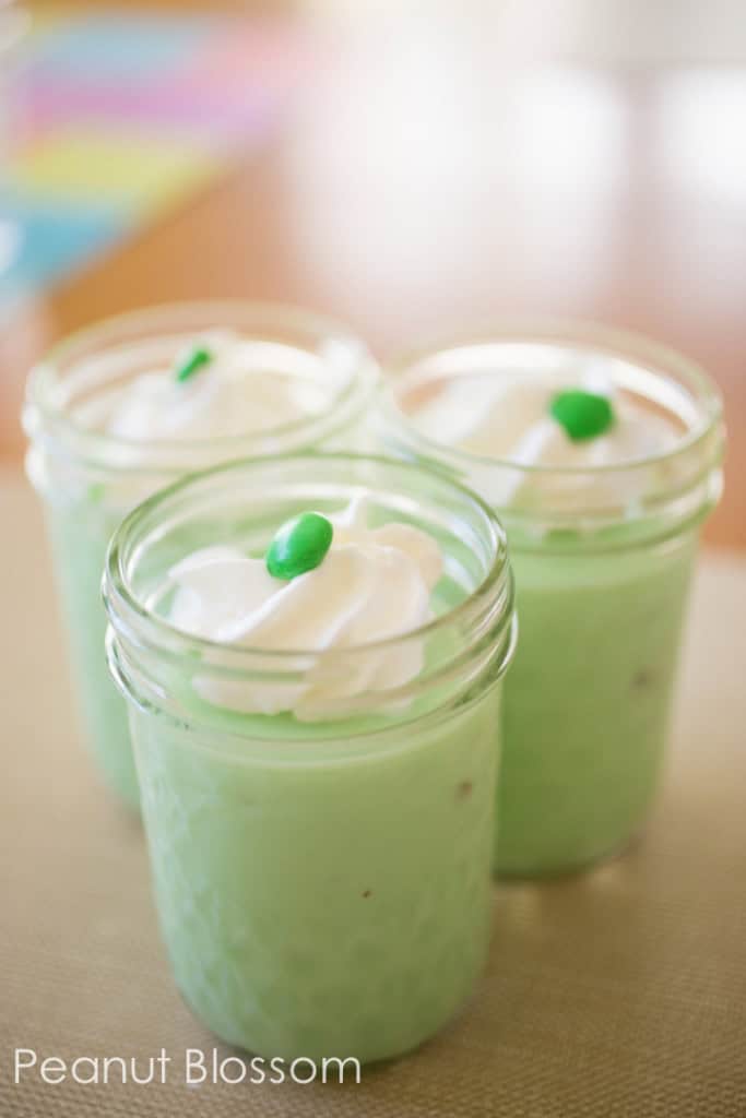 Mason jars filled with pistachio pudding have a dollop of whipped cream and a green M&M candy on top.