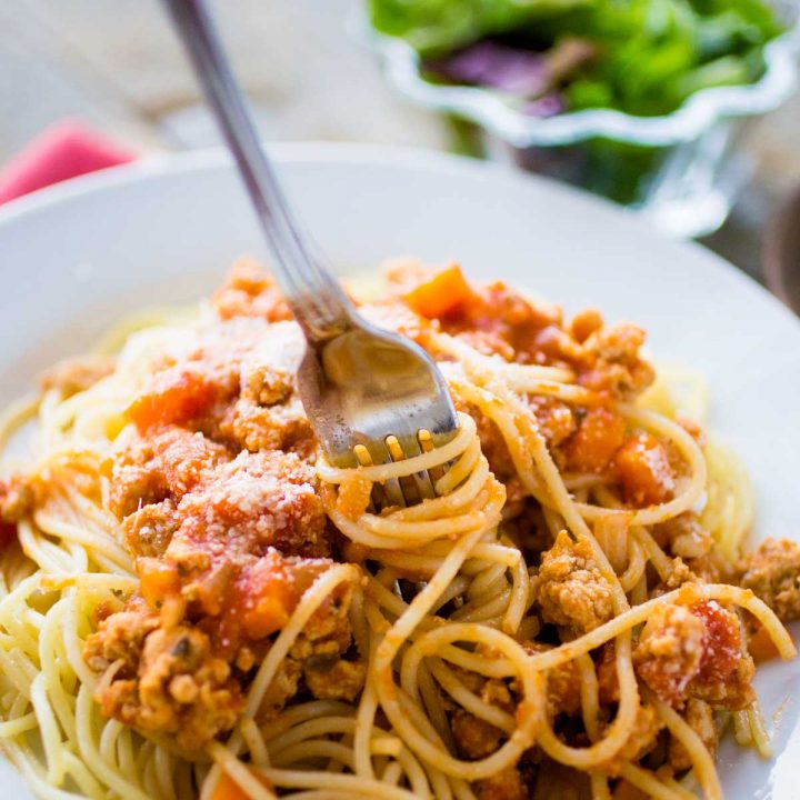 A plate of spaghetti with chicken bolognese sauce has a fork twirling the noodles.