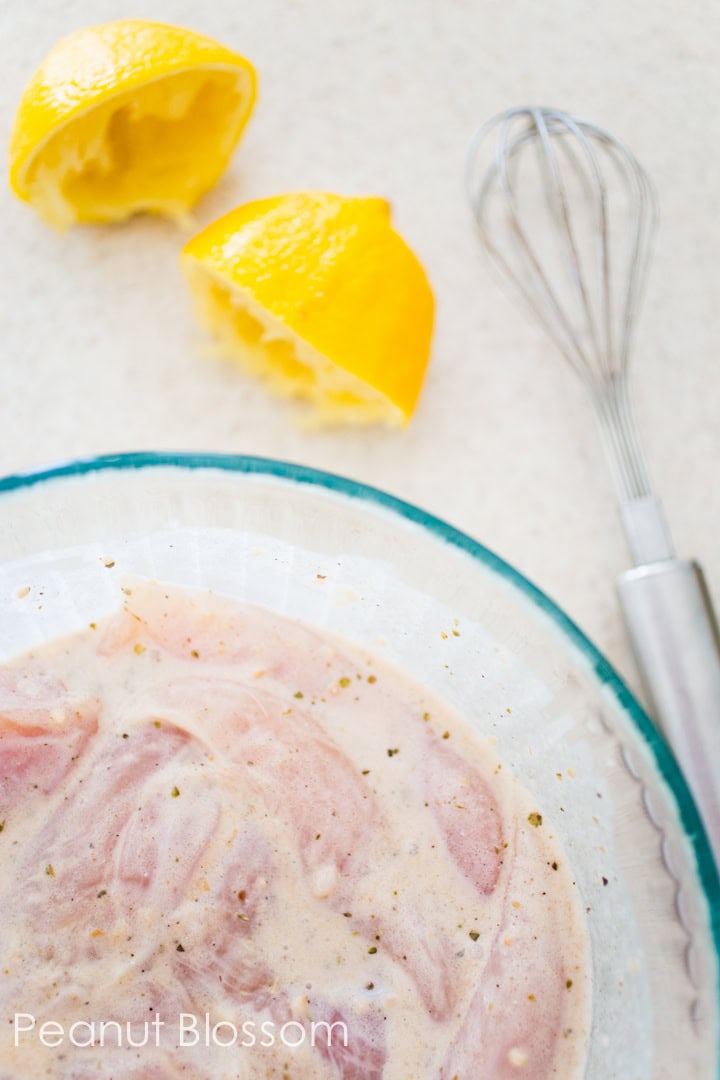 The chicken is marinating in a large mixing bowl with a whisk and squeezed lemon halves on the counter next to it.