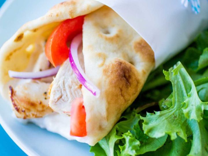 A pita is wrapped around chicken gyro meat with fresh tomatoes and slices of red onion. The sandwich sits next to some fresh lettuce.