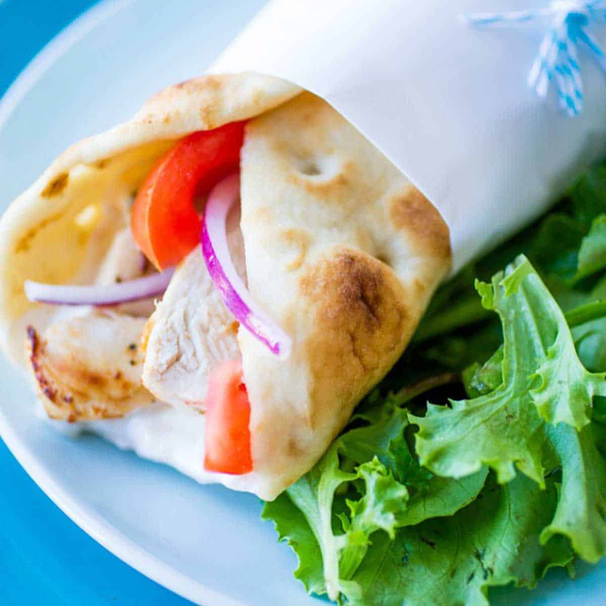 A pita is wrapped around chicken gyro meat with fresh tomatoes and slices of red onion. The sandwich sits next to some fresh lettuce.