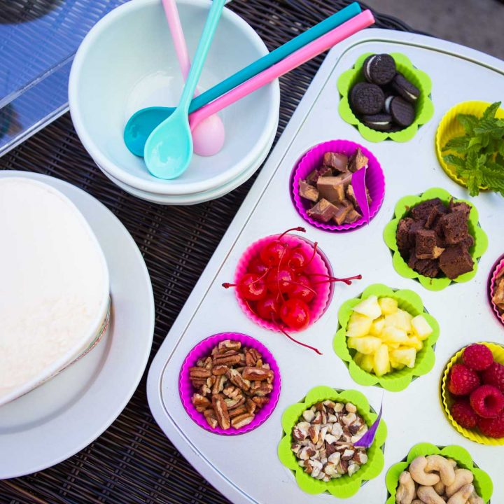 A muffin tin is filled with all different sundae bar toppings including chopped nuts, brownies, cherries, cookies, and fresh mint.
