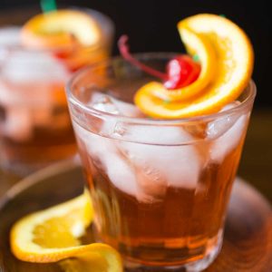 A glass of brandy old fashioned sweet has lots of ice and a maraschino cherry with orange twist for garnish.