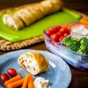 A serving of the chicken cheese roll up sits on a plate next to fresh carrots, tomatoes, and a veggie tray with dip.