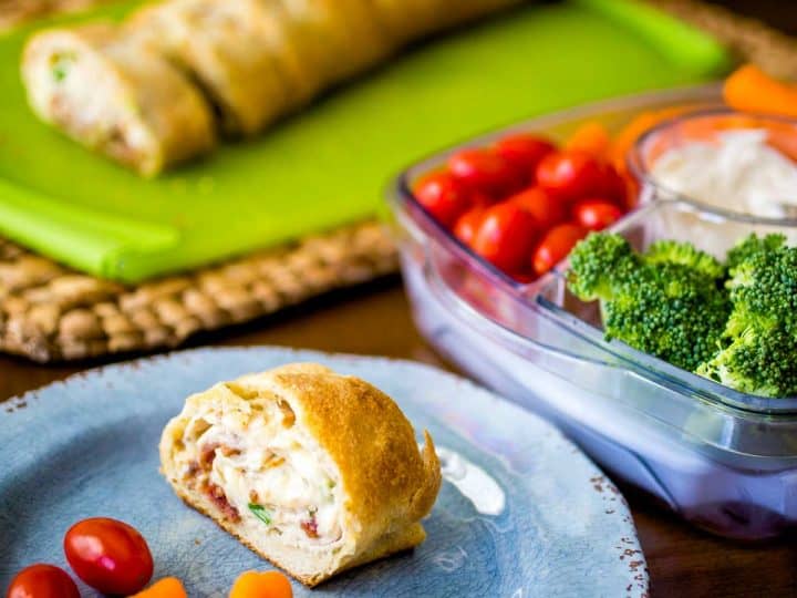 A serving of the chicken cheese roll up sits on a plate next to fresh carrots, tomatoes, and a veggie tray with dip.