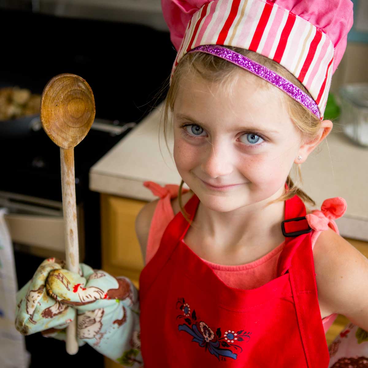A young girl has an apron, oven mitt, cooking spoon, and chef hat.