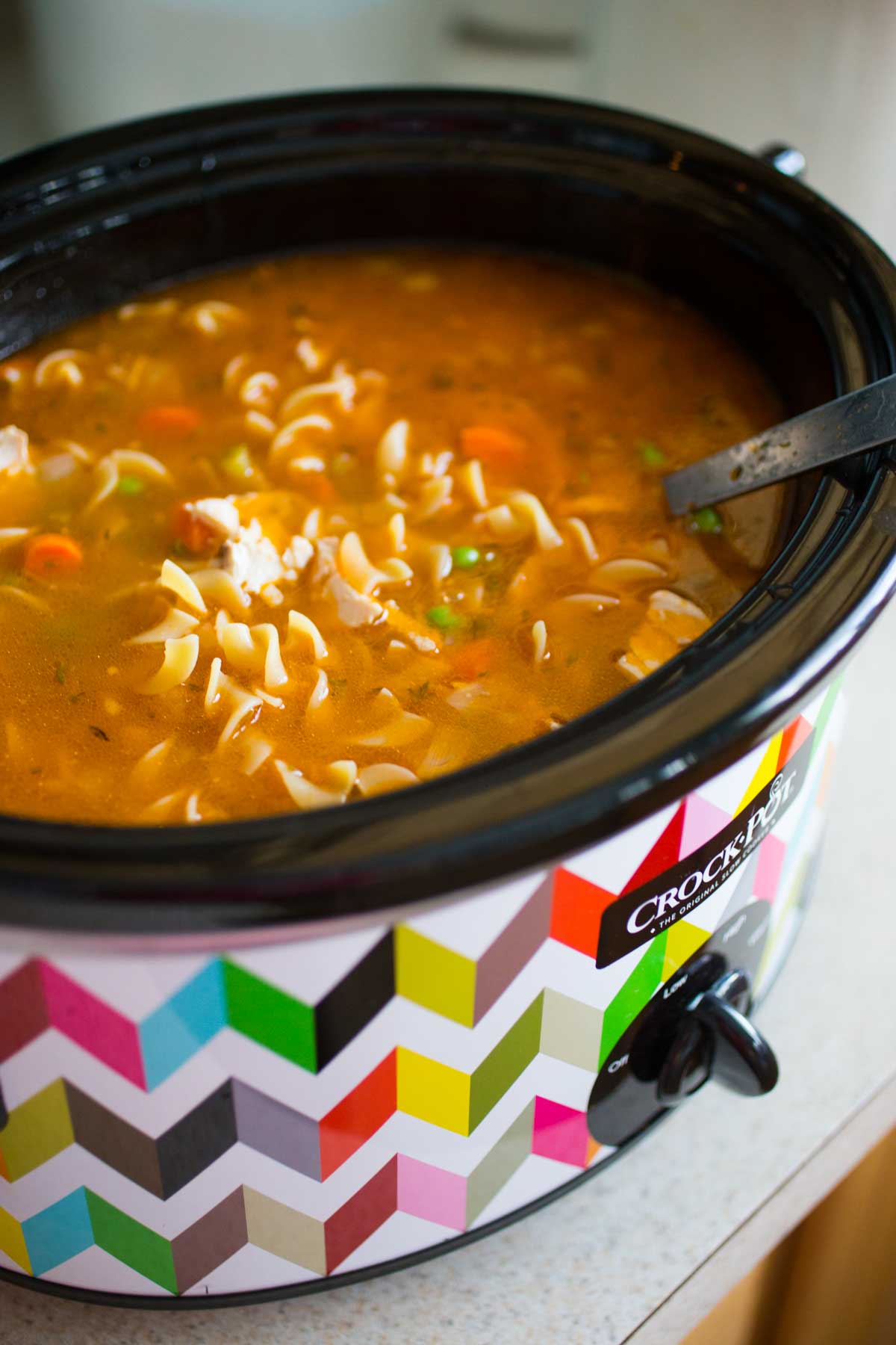 A colorful Crockpot is filled with a tomato-based chicken soup with pasta.