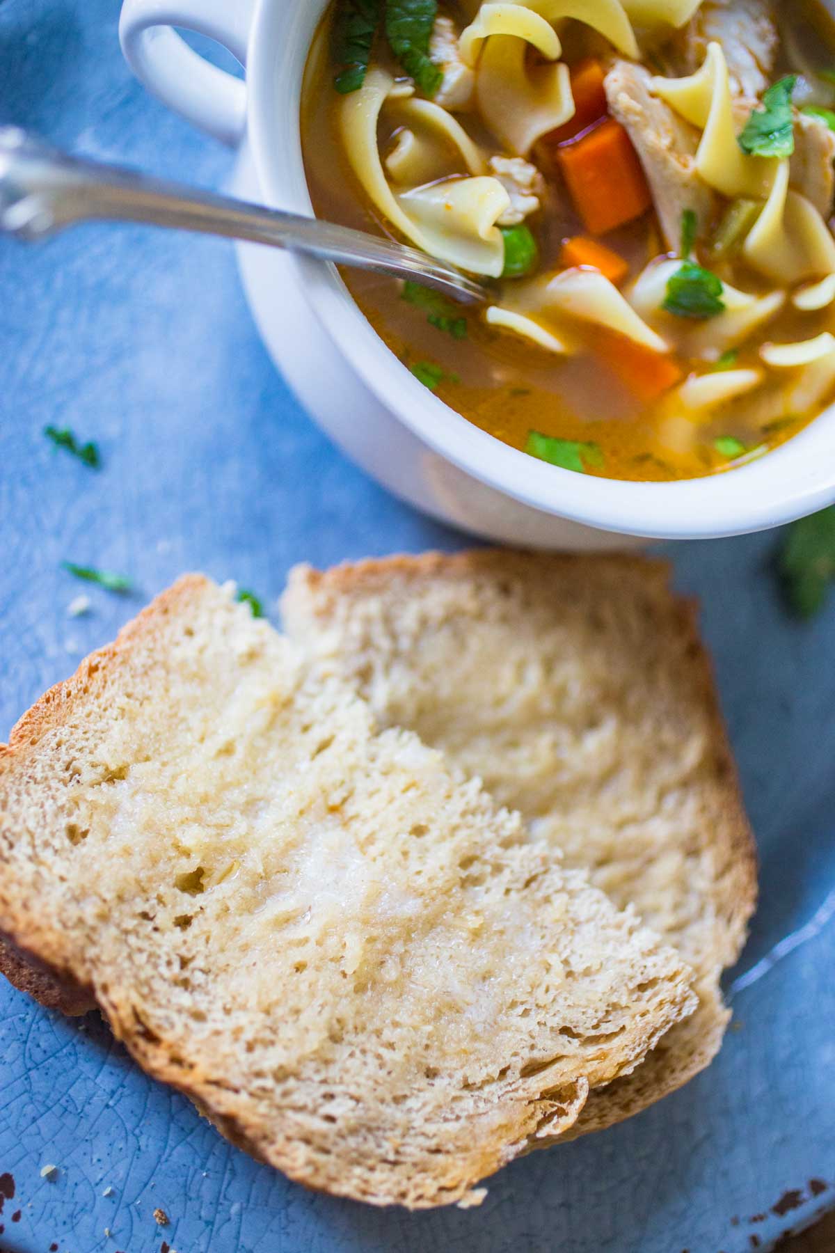A bowl of chicken soup sits on a blue plate next to a buttered slice of fresh bread.
