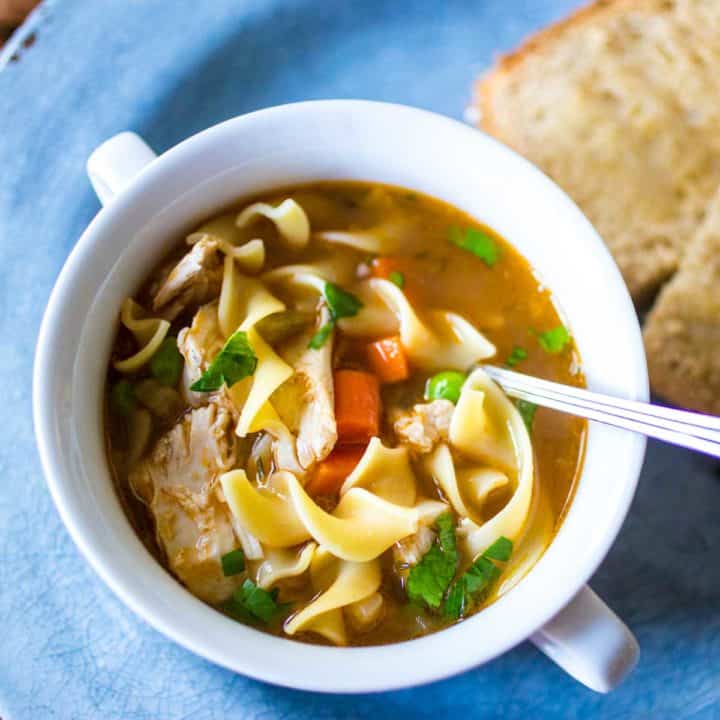 A white bowl of chicken noodle soup sits on a blue plate. There are wavy egg noodles and fresh parsley peeking out of the broth.