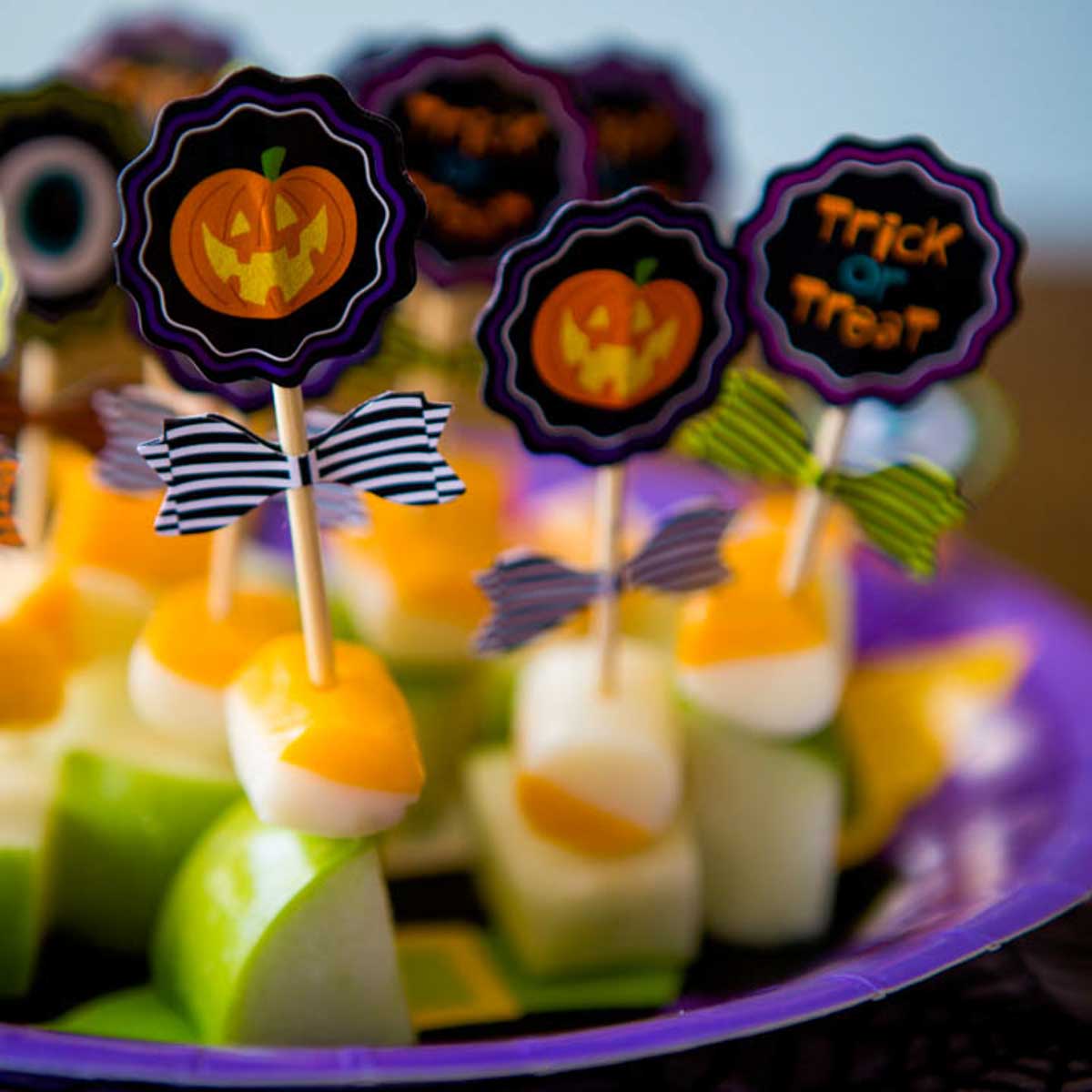 A purple plate has festive skewers of fresh green apples and striped orange cheese chunks with a Halloween pumpkin pick.