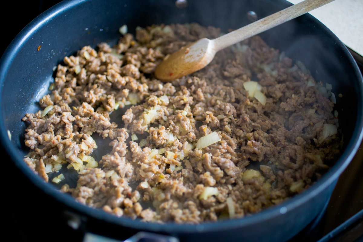 The skillet has browned bulk sausage with chopped onion being stirred in with a spoon.
