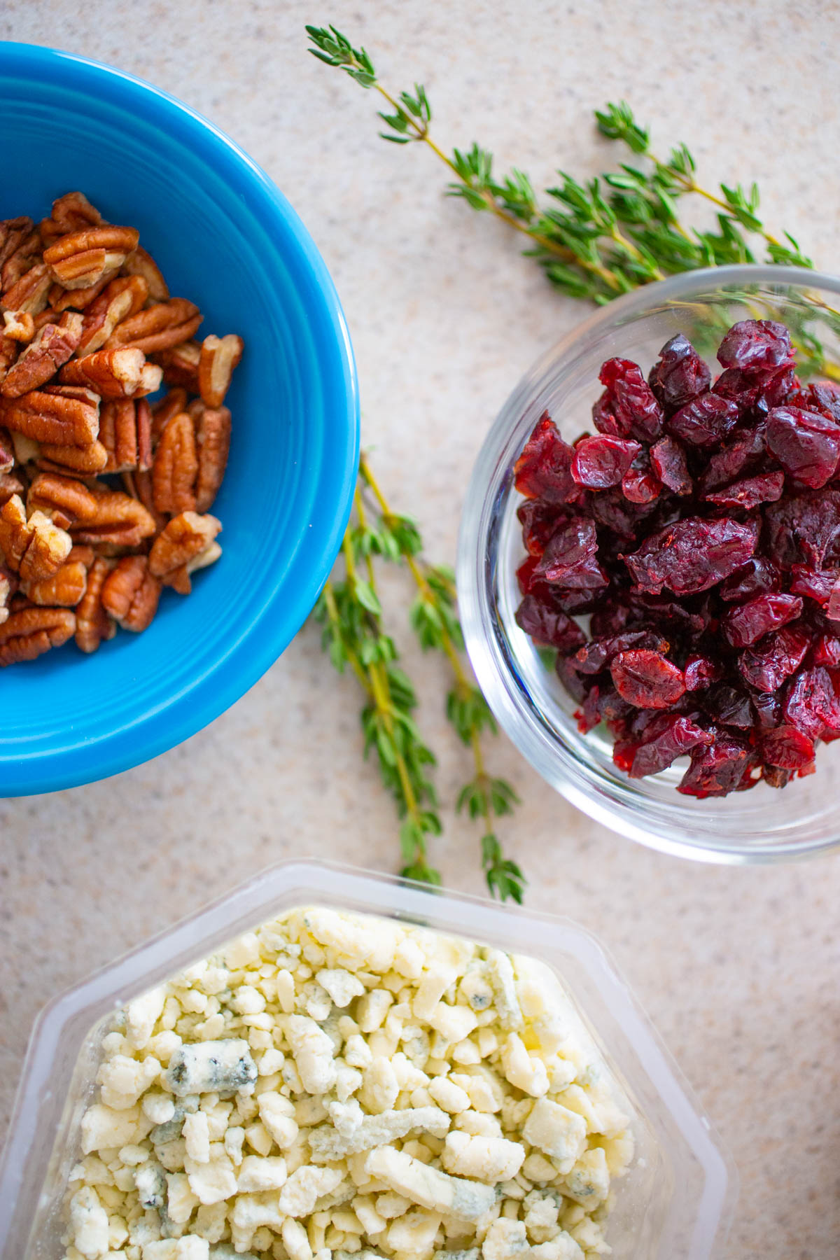 The pecans, cranberries, and crumbled blue cheese are on the counter next to fresh thyme.