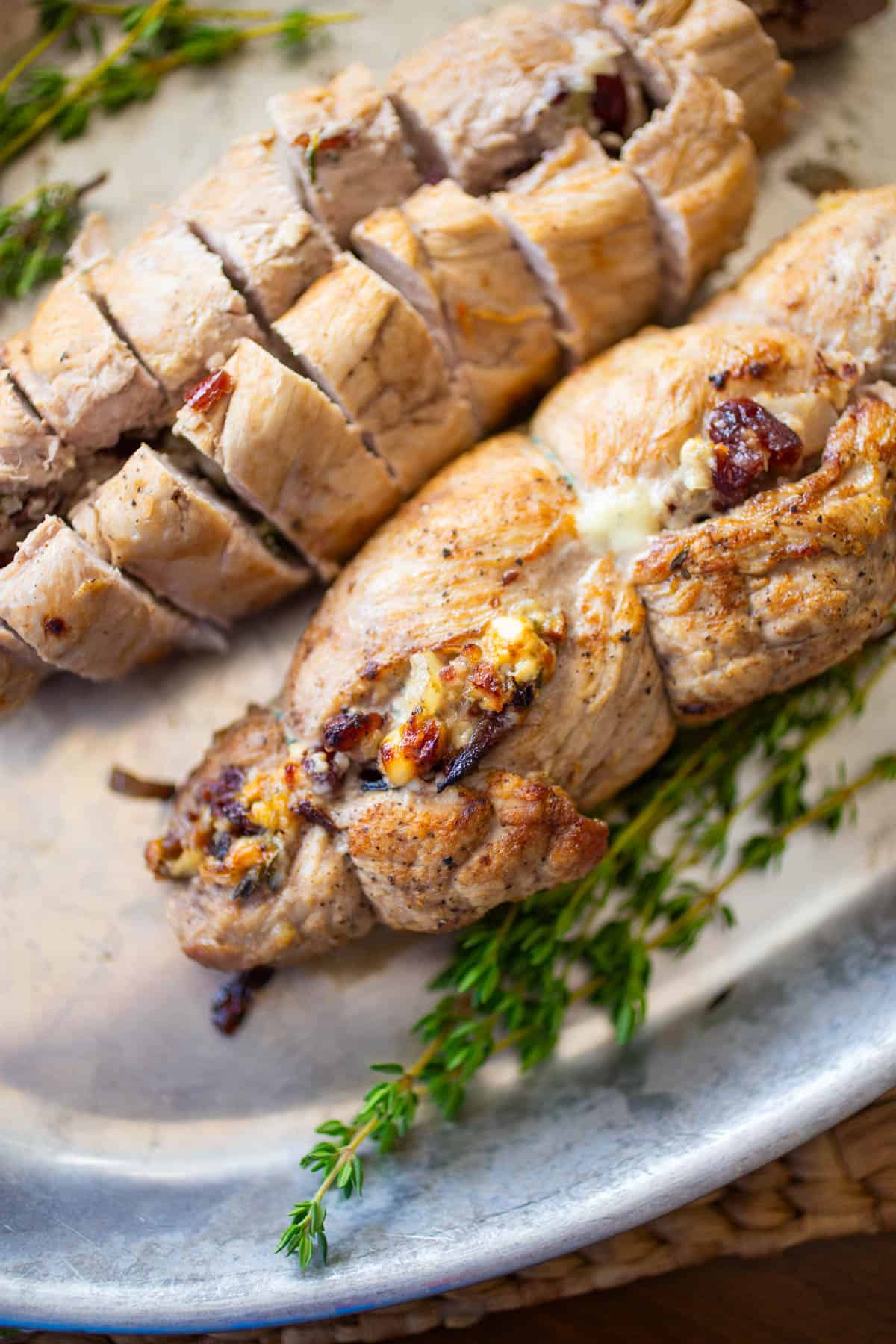 The stuffed tenderloins have been baked and are golden brown. They are on a metal serving pan with fresh thyme on the side.