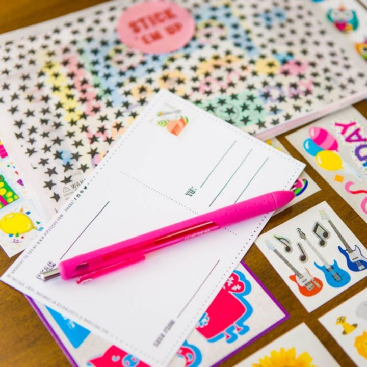 A blank notecard, pink pen, and colorful stickers and stamps are displayed on a table.