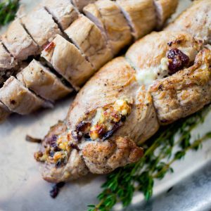 A stuffed pork tenderloin has cranberries and blue cheese peeking out of the roll. A bunch of fresh thyme is on the platter.