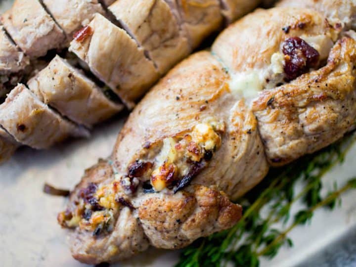 A stuffed pork tenderloin has cranberries and blue cheese peeking out of the roll. A bunch of fresh thyme is on the platter.