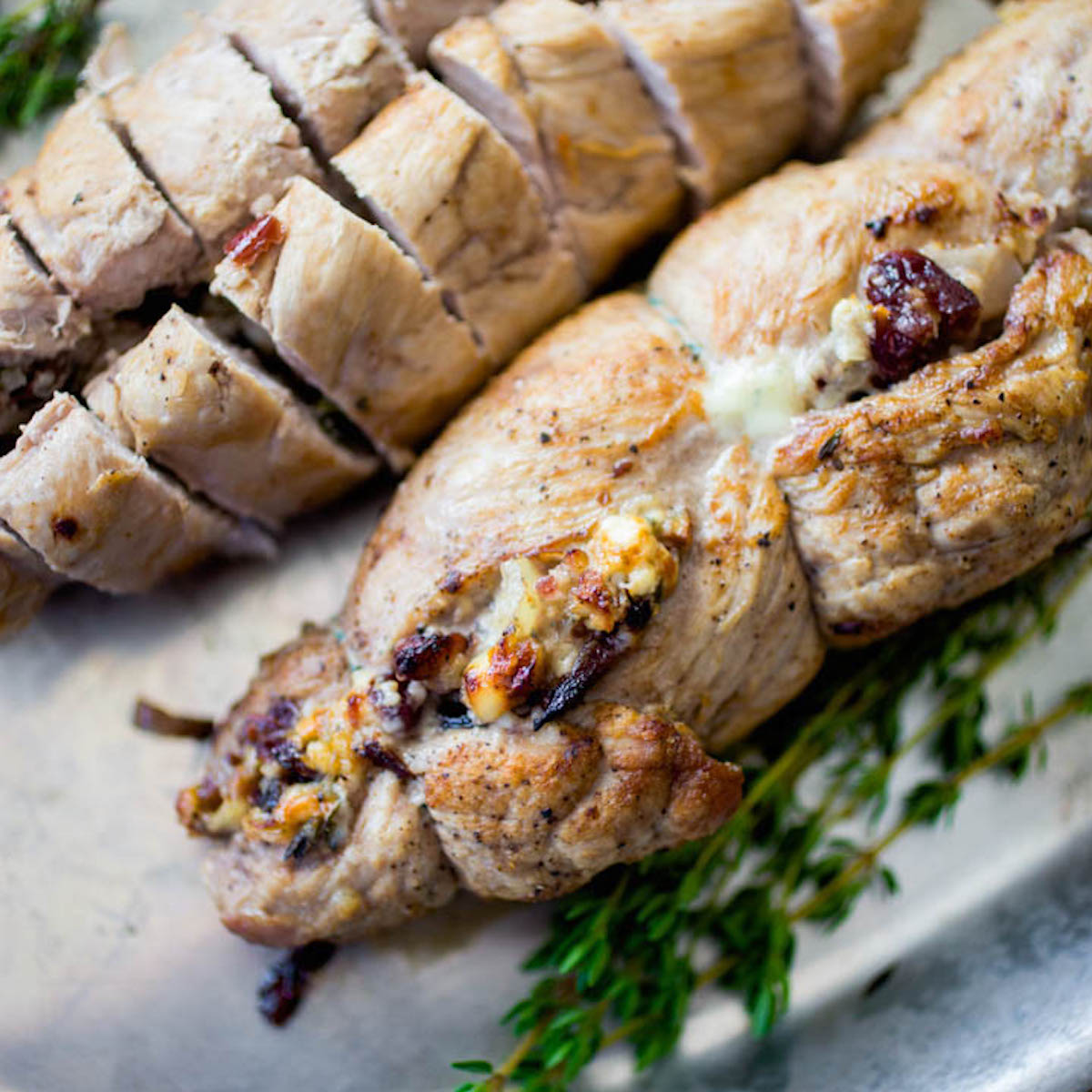 A stuffed pork tenderloin has cranberries and blue cheese peeking out of the roll. A bunch of fresh thyme is on the platter.