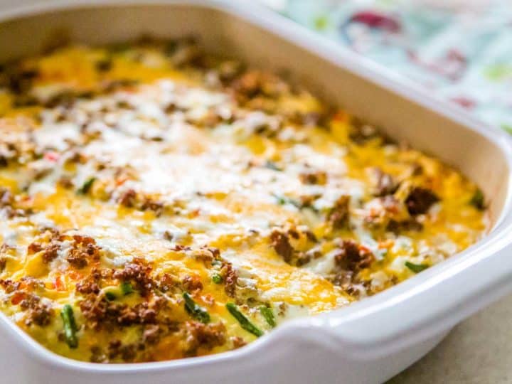 A white baking dish has a cheesy sausage and egg casserole with green veggies mixed in.