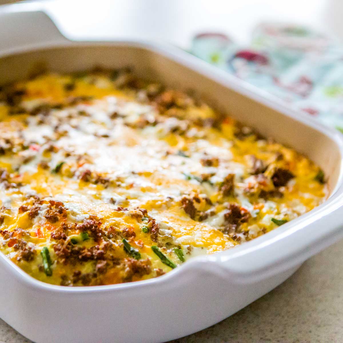 A white baking dish has a cheesy sausage and egg casserole with green veggies mixed in.