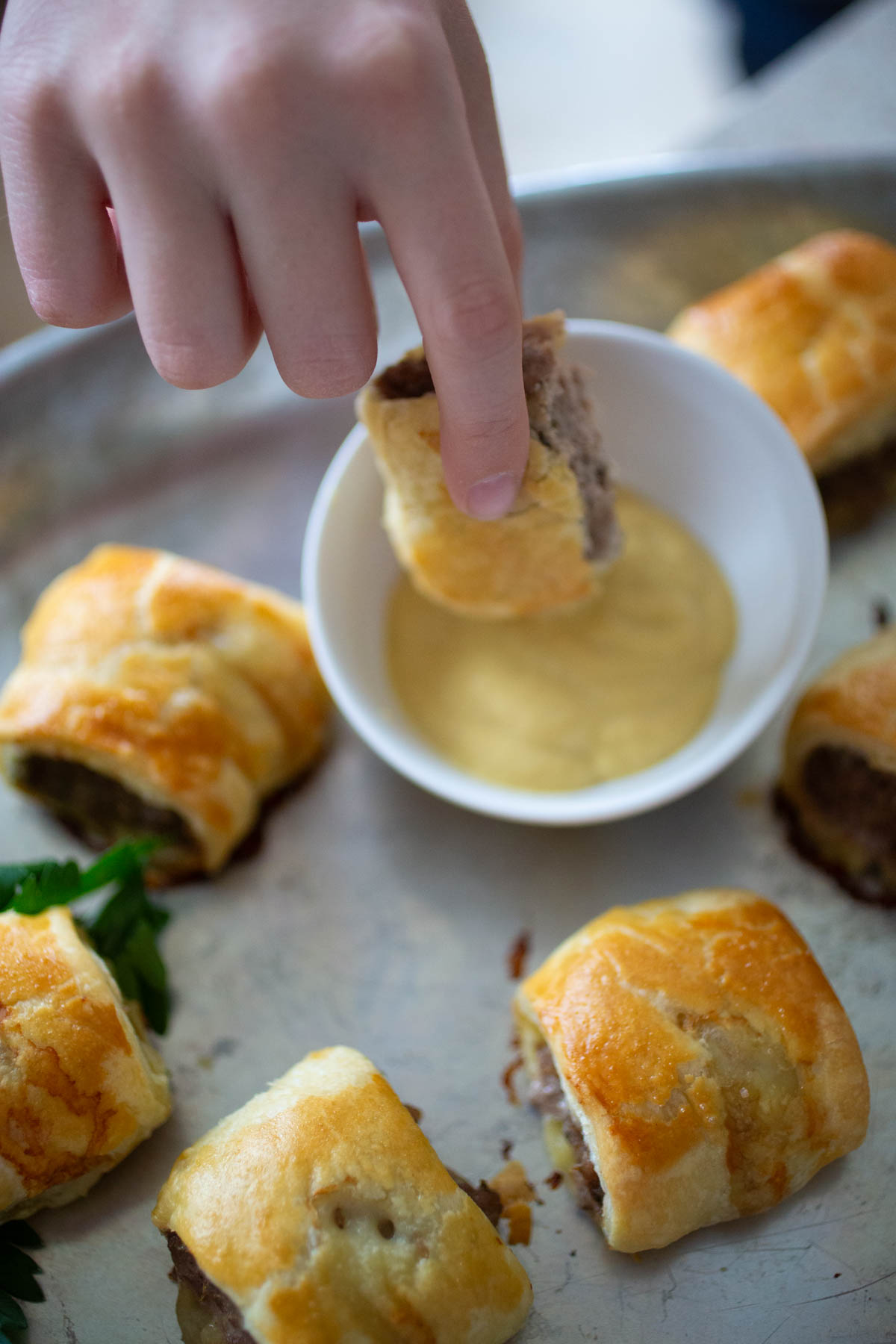 A hand is dipping one of the sausage rolls into a bowl of mustard.