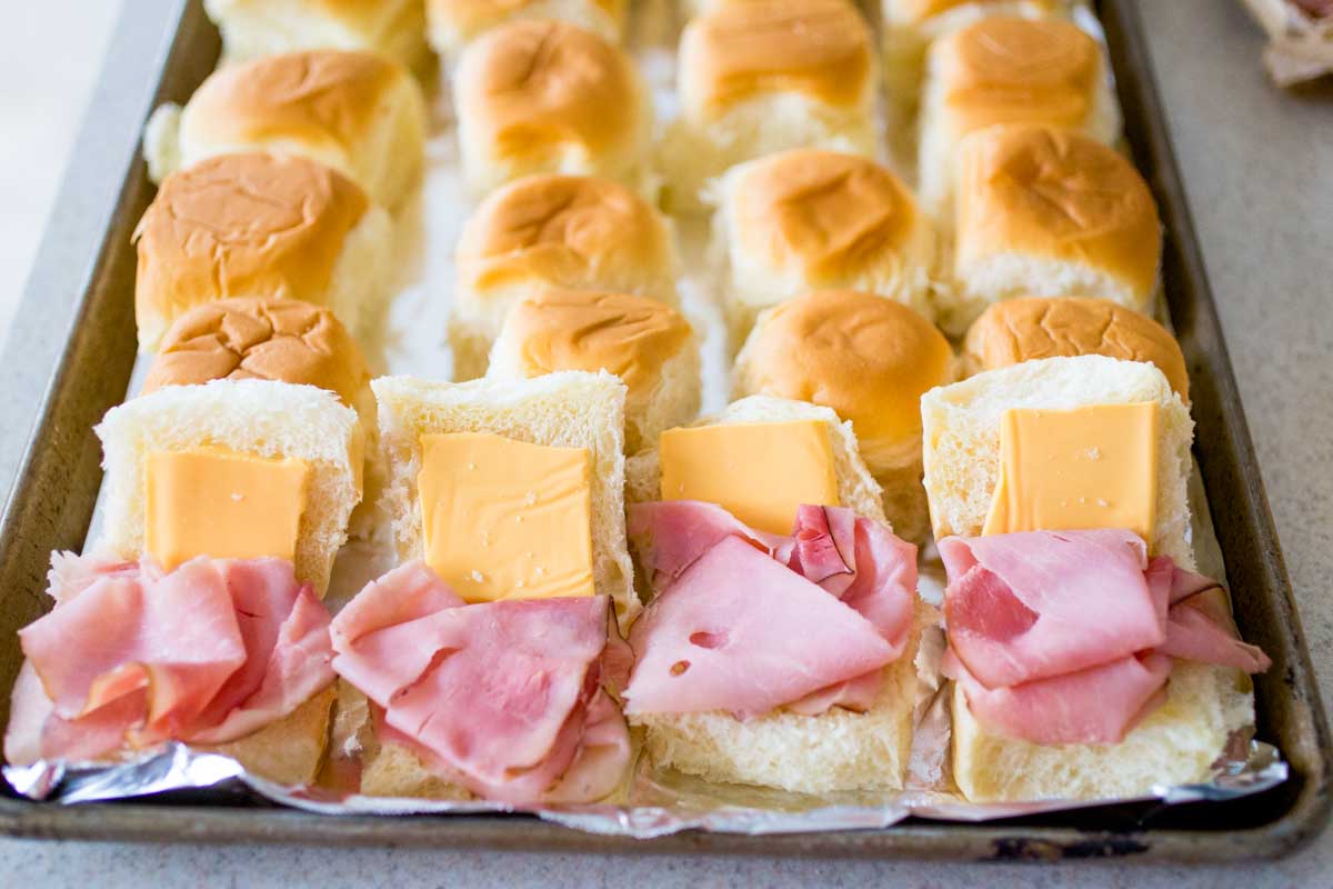 The ham and cheese have been added to the slider buns on a baking pan.