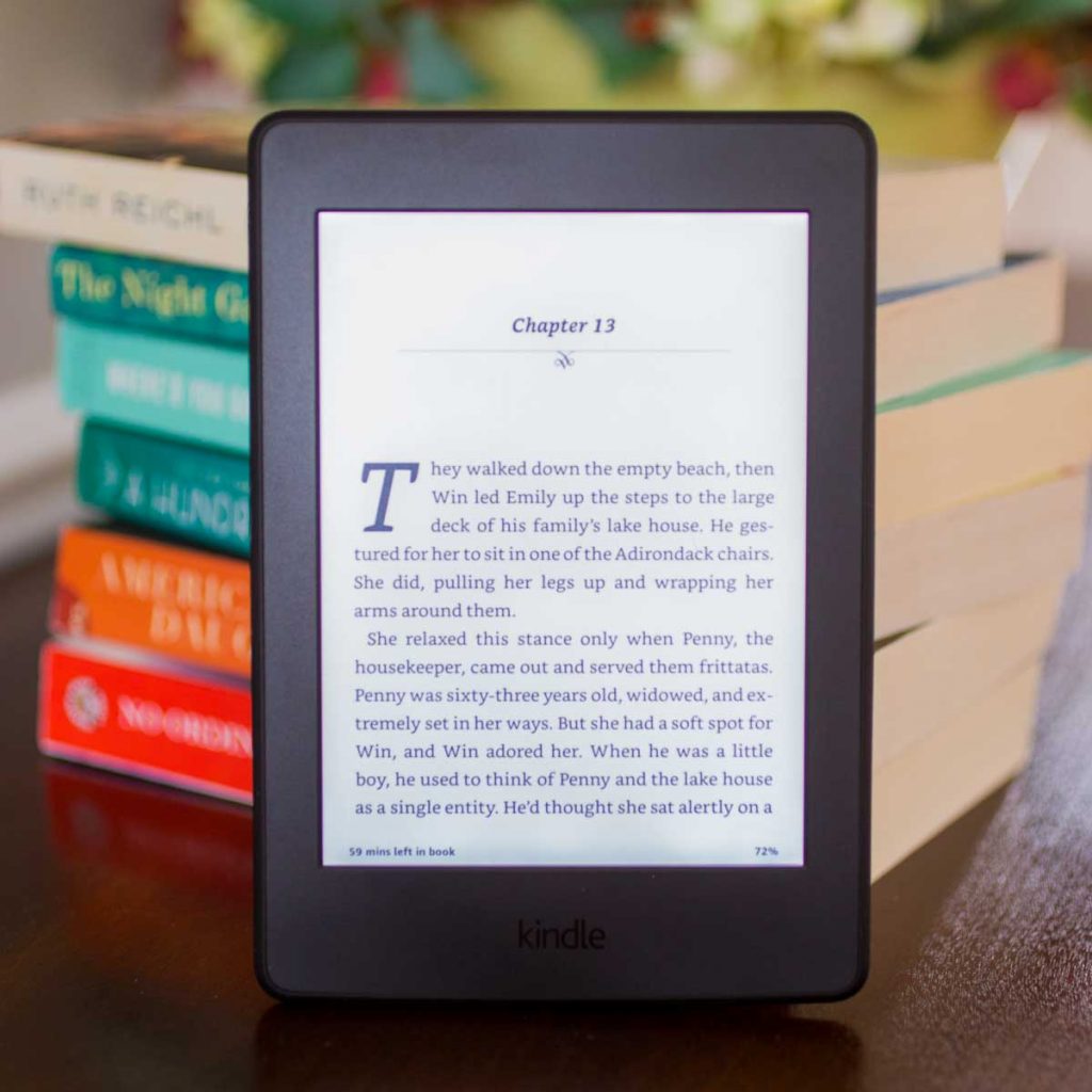 A Kindle eReader sits in front of a stack of paper books.