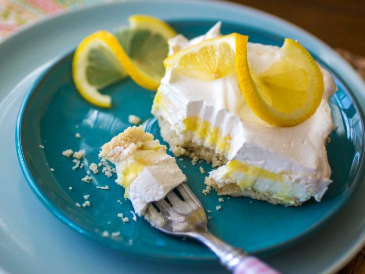 A slice of layered lemon torte sits on a blue plate. You can see the yellow lemon layer, a white whipped topping layer, and the graham cracker crust. A twirled slice of lemon sits on top for garnish.
