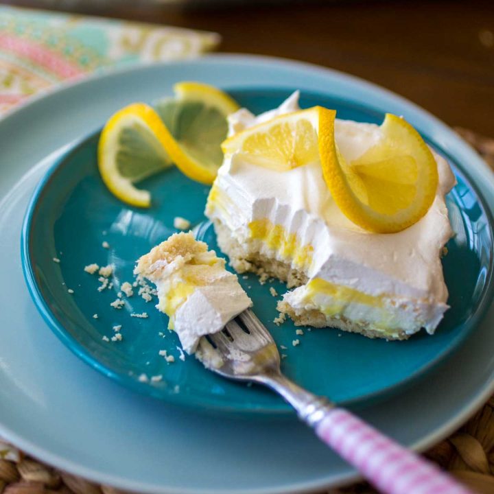 A slice of layered lemon torte sits on a blue plate. You can see the yellow lemon layer, a white whipped topping layer, and the graham cracker crust. A twirled slice of lemon sits on top for garnish.