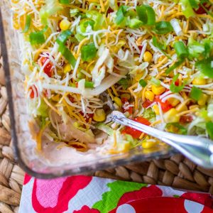 A container of homemade taco dip has a spicy cream cheese filling and is topped with shredded cheese and lettuce. A serving spoon has scooped a portion out.