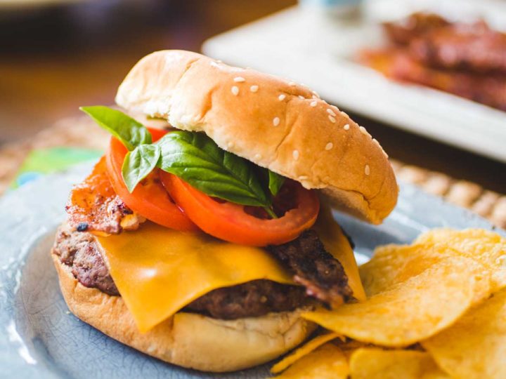 A bacon cheddar burger has layers of fresh tomato and fresh basil and sits next to a serving of potato chips.